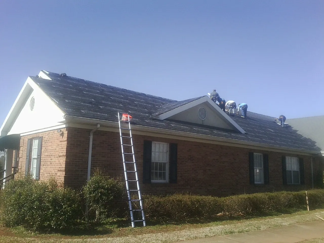 Crew tearing shingles off a brick ranch roof with a ladder set — Metal Roof Repair work by our crew in Brogue