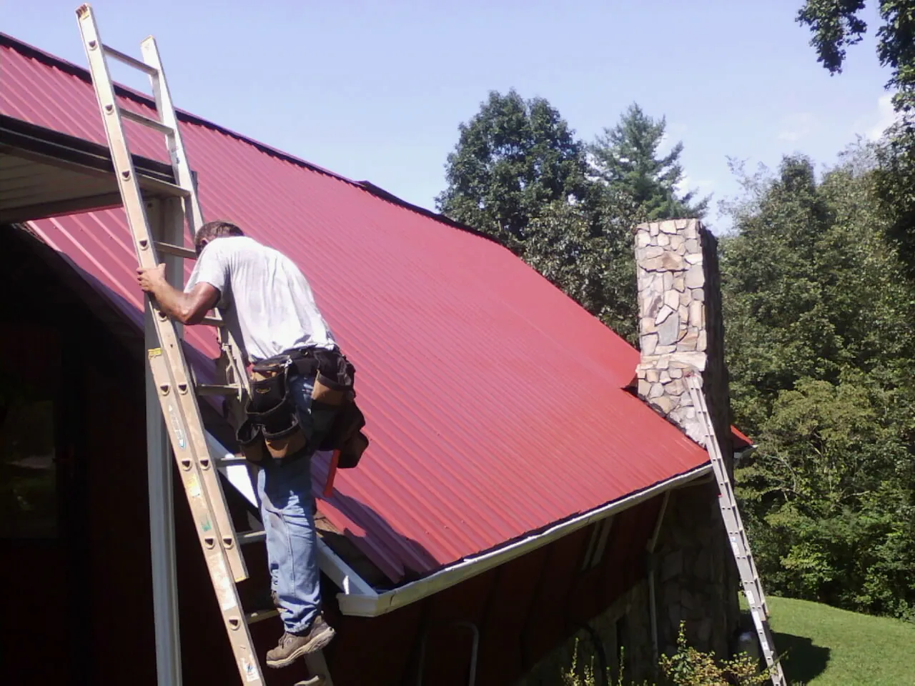 Roofer on a ladder beside a red metal roof and stone chimney — Soffit & Fascia Repair by Roofing Brothers in Brogue